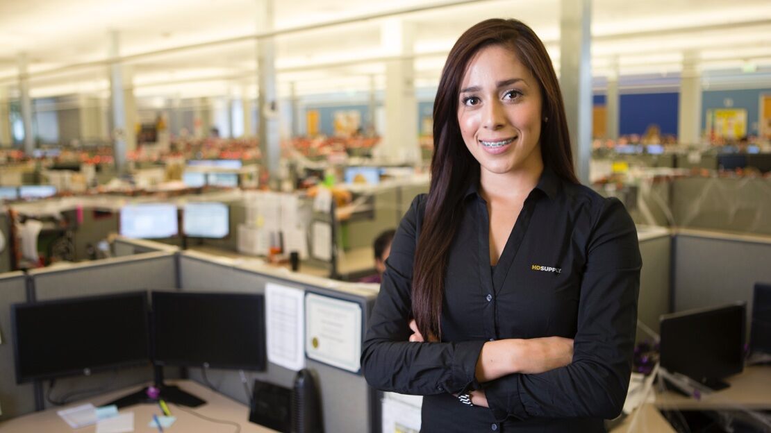 Young woman in an HD Supply dress shirts stands among cubicles with her arms crossed