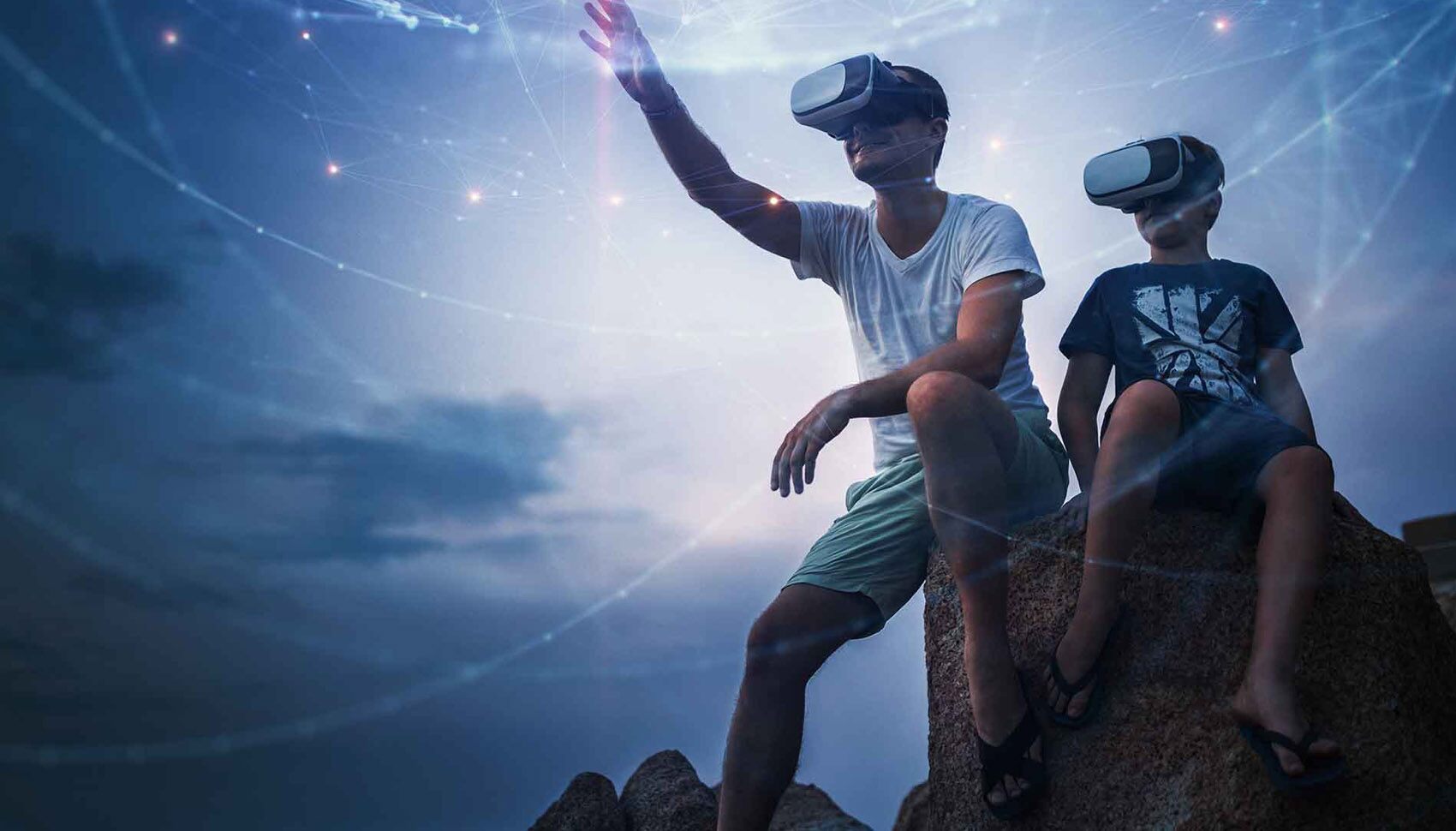 A dad and his young son are seated on a rock looking out with virtual reality googles on