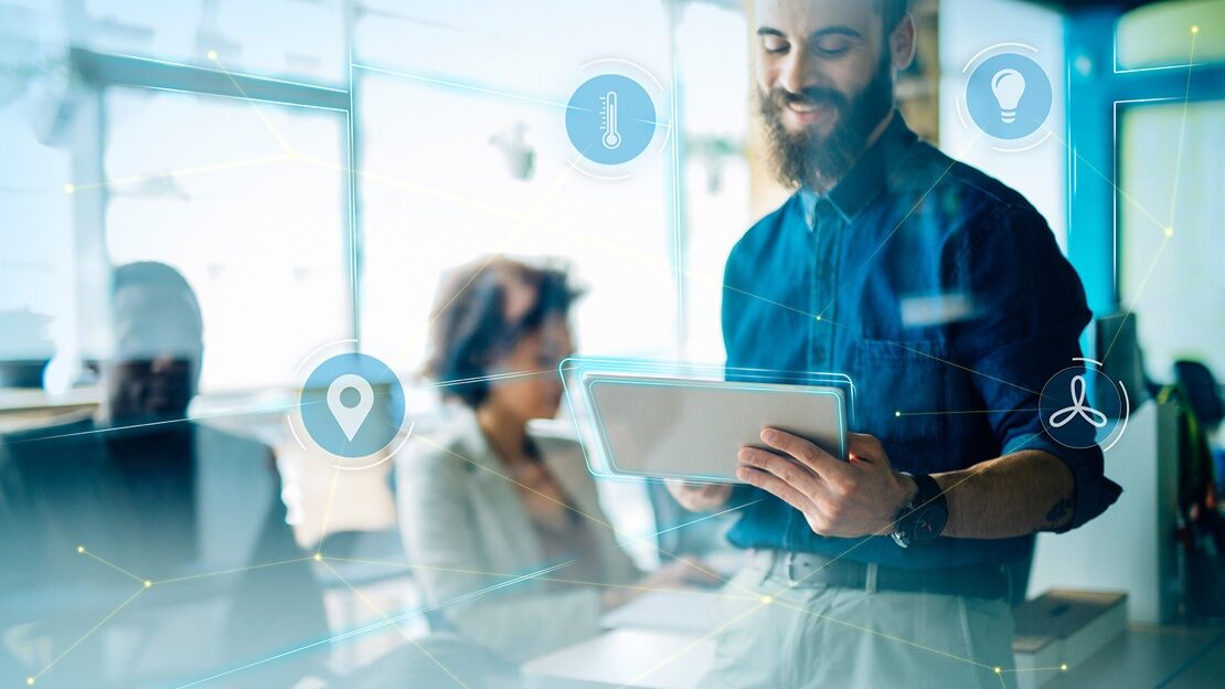 A bearded young man leans on a desk, focused on an iPad with work colleagues seated in the background