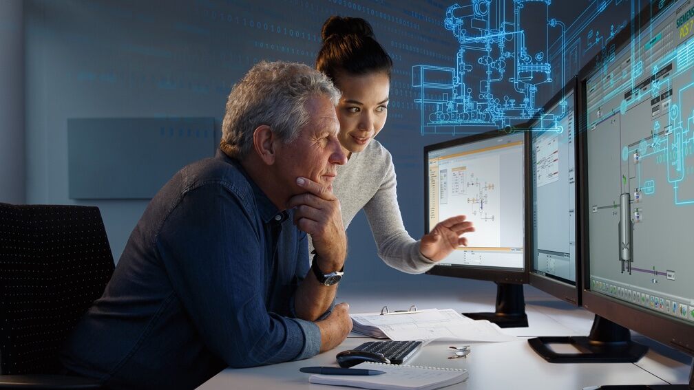 An older man and a younger woman at a desk looking at 3 PC monitors
