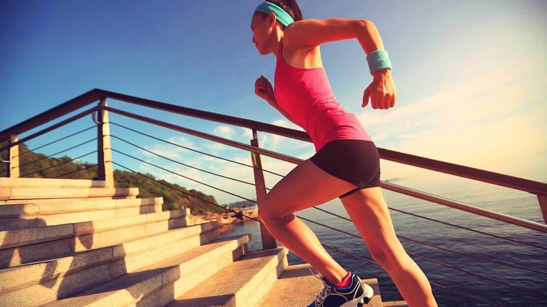 A young woman in exercise clothing runs up concrete steps with the sea in the background