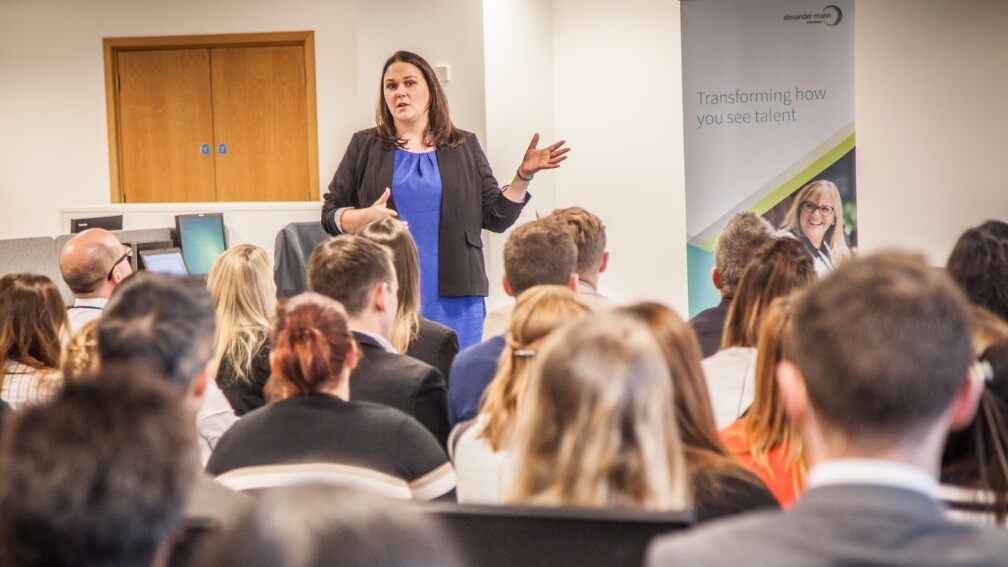 An Alexander Mann Solution’s female executive stands among a group of employees who are seated in a conference room