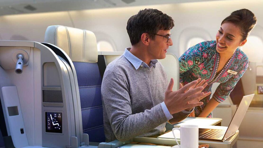 A male passenger sits in a first-class seat talking to a smiling female Malaysia Airlines flight attendant