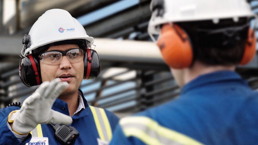 Two men in blue worker uniforms walk towards a Nexen Energy power plant