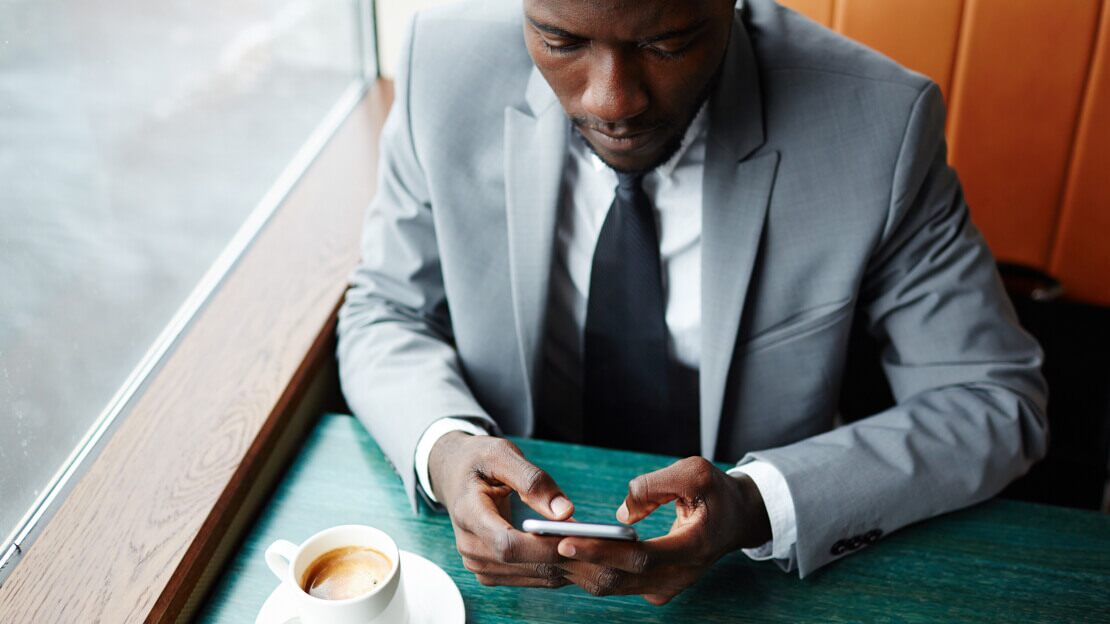 A businessman in a suit is sitting at a table with a cup of coffee, working on his mobile phone