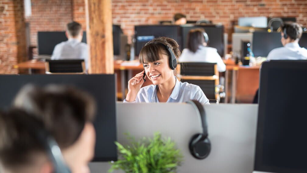 Focal point is on a young smiling woman with a head set on working in a modern call centre surrounded by co-workers