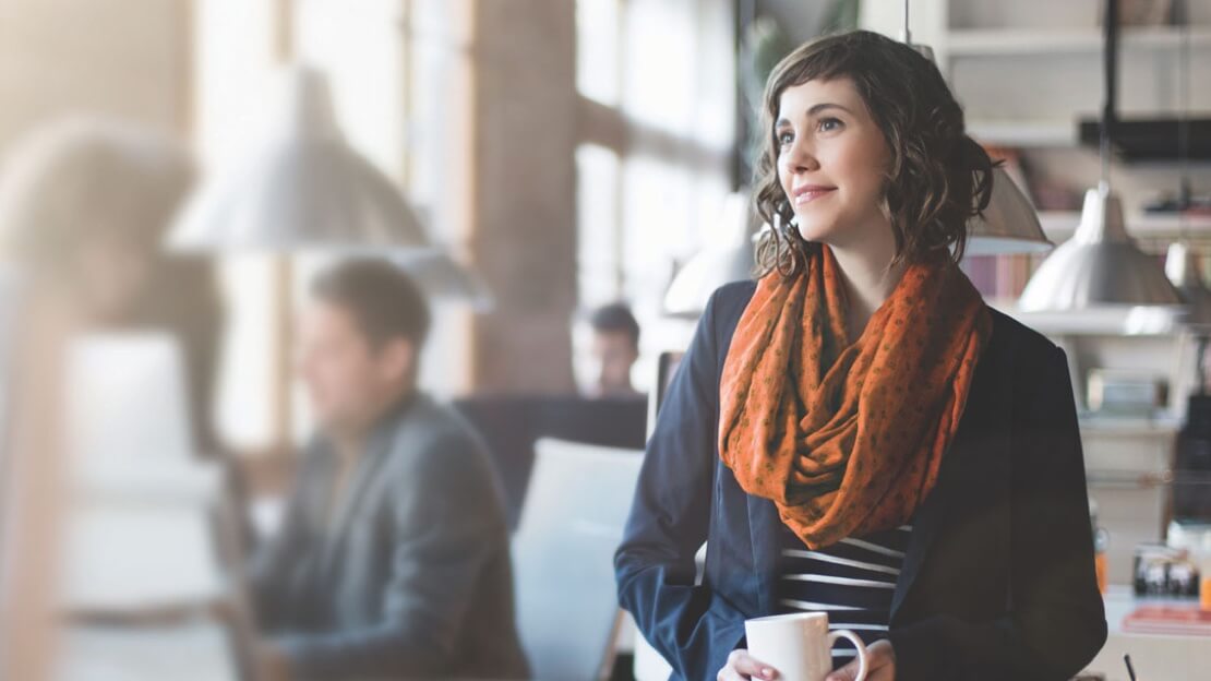 A young woman with long dark hair sits in an office, looks off as she holds a cup of coffee
