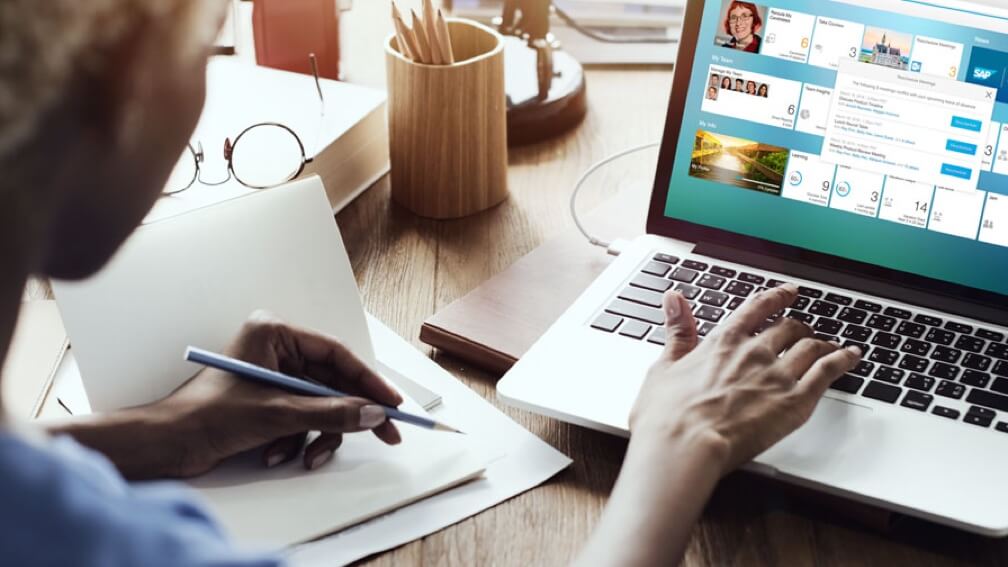  Looking over a woman’s shoulder at an open laptop working it with her right hand while holding a pencil in her left hand